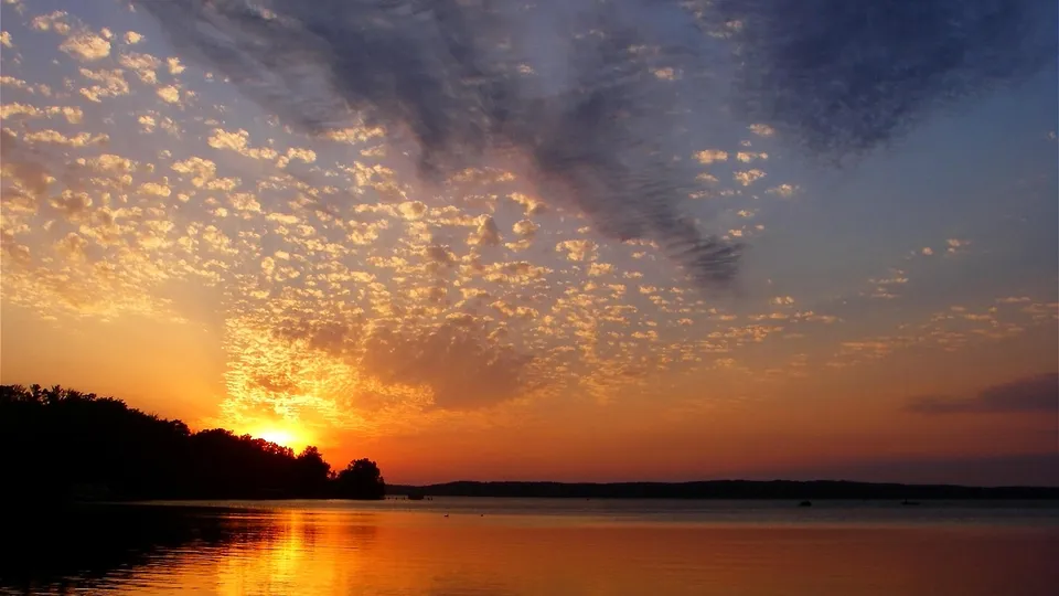 A warm orange sunset glow behind a silhouetted treeline, reflected on the still water of a lake.