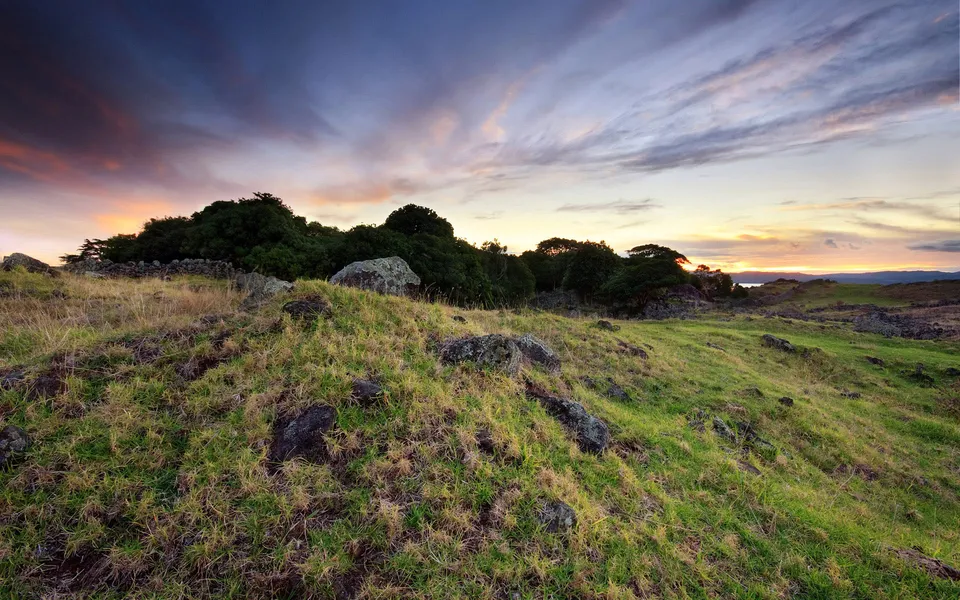 A dramatic sunset with colorful clouds over a rolling green hillside dotted with rocks and trees.