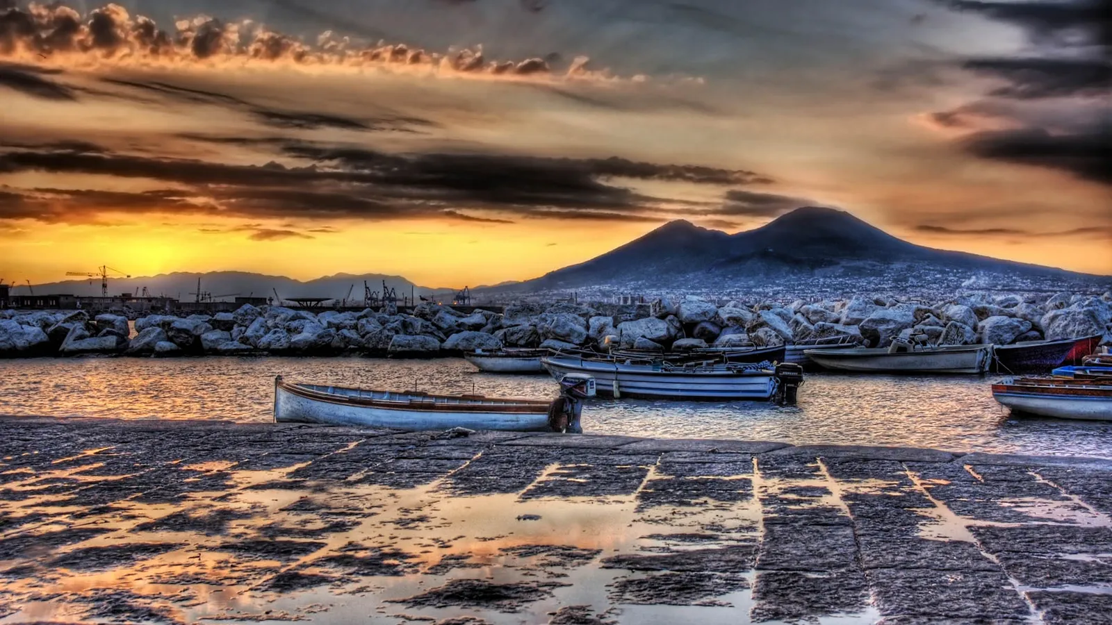 A dramatic sunset over a harbor with boats docked, with Mount Vesuvius in the background.
