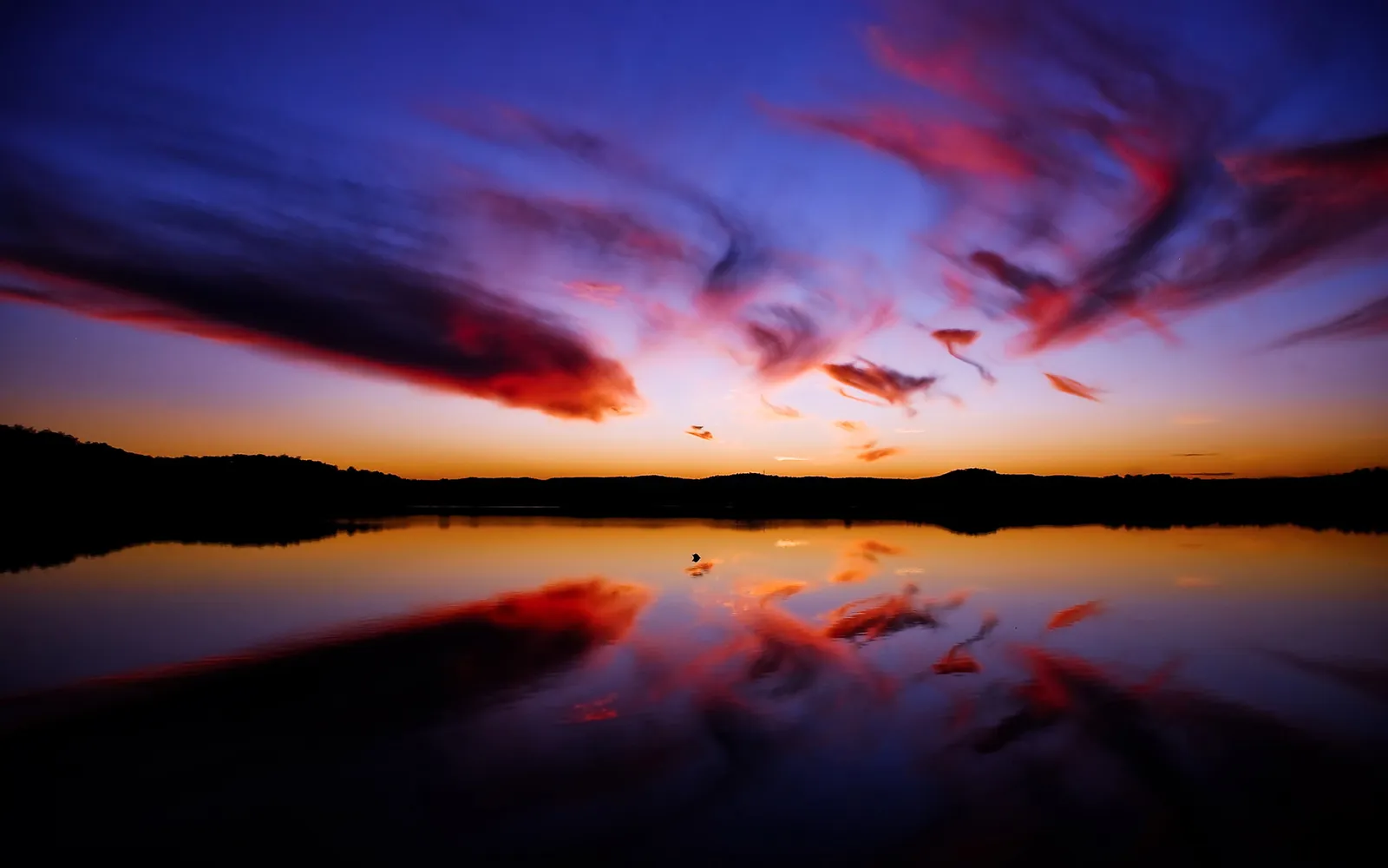 A vibrant sunset with red and purple clouds reflected in a calm lake, silhouetting a distant hill.