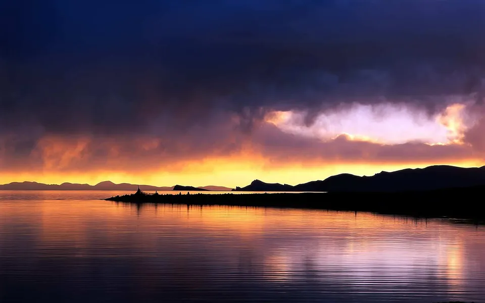 A dramatic sunset with vibrant orange and purple clouds reflects on a calm lake with silhouetted mountains in the distance.