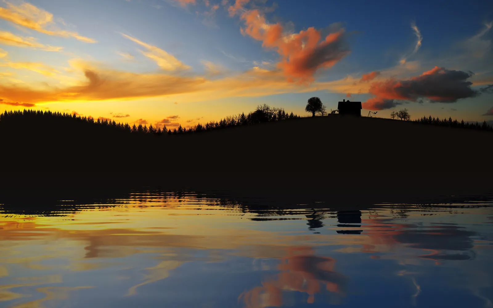 A silhouetted hill with trees and a house is reflected in a calm lake during a vibrant sunset.
