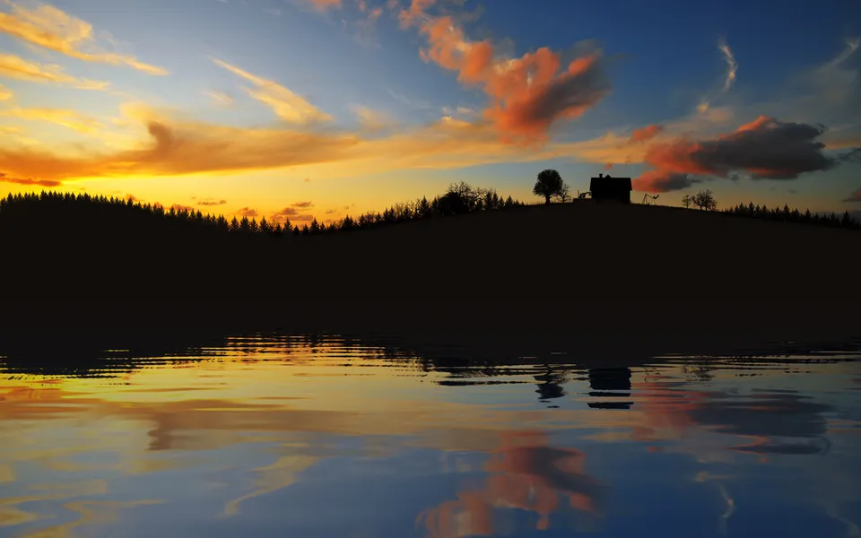 A silhouetted hill with trees and a house is reflected in a calm lake during a vibrant sunset.
