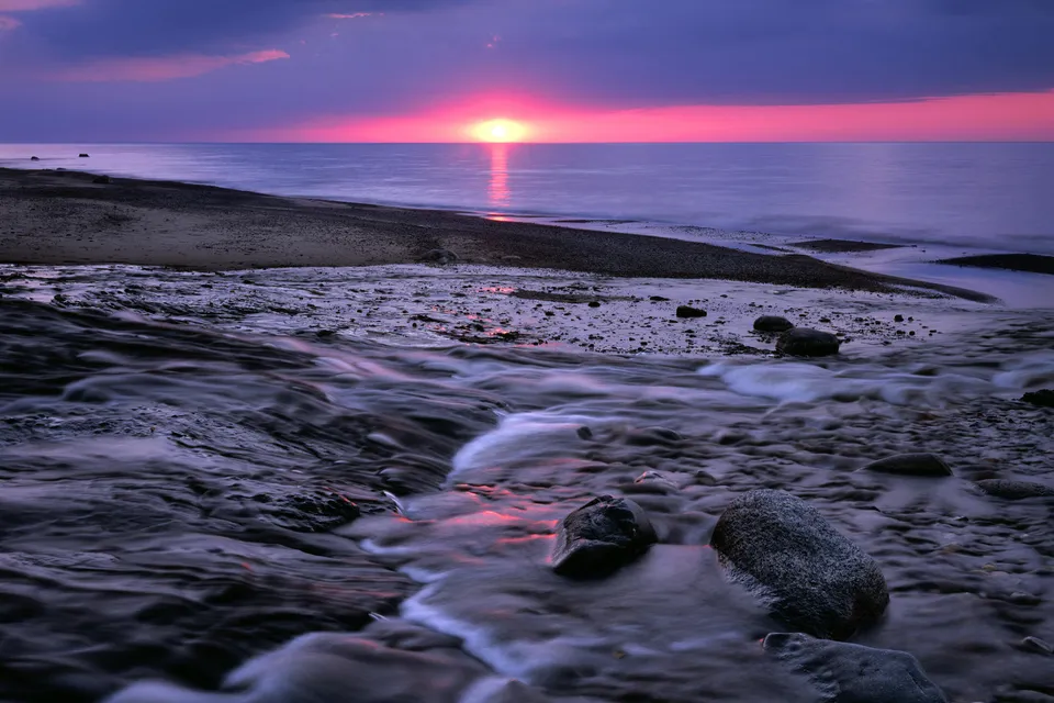 A vibrant sunset paints the sky with pink and purple hues over a calm lake, with a small stream flowing into the water.
