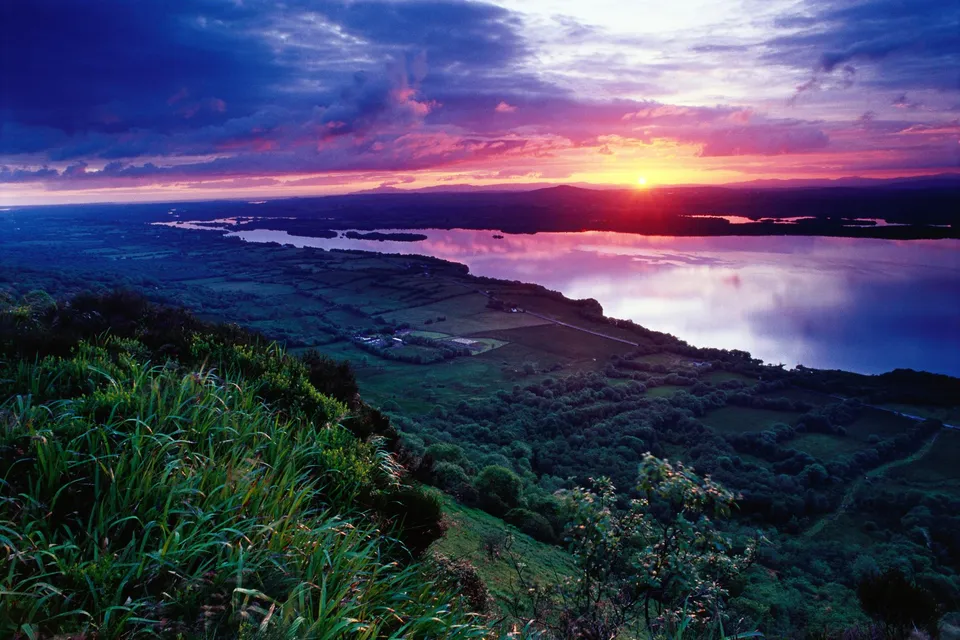 A dramatic sunset over a wide lake, with colorful clouds reflecting in the water and a green hillside in the foreground.