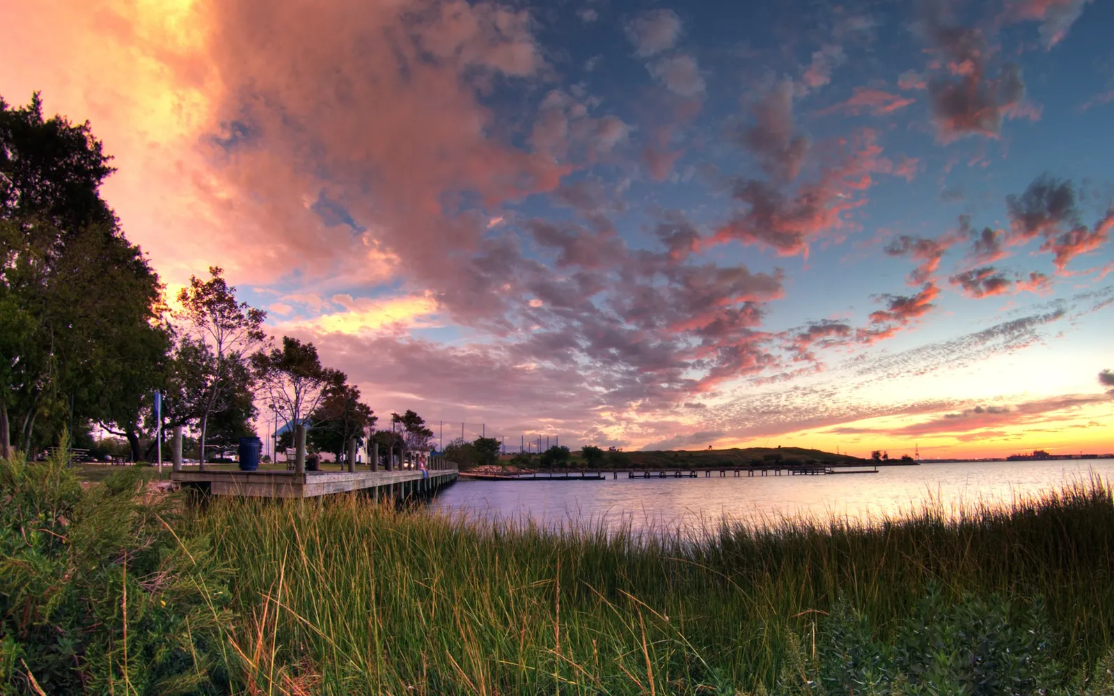 A sunset over calm coastal waters, with a pier, marsh grasses, and trees in the foreground.