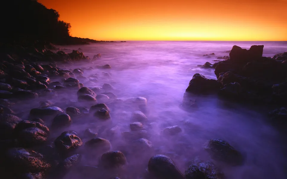 A rocky shoreline is bathed in purple mist as the sun sets with an orange glow on the horizon.