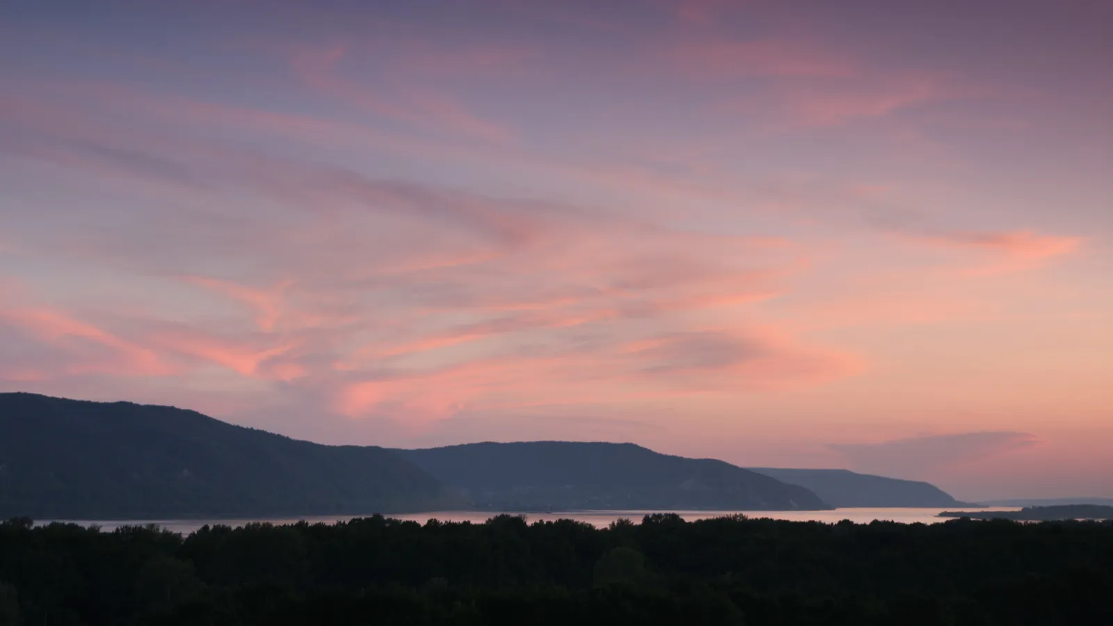 A serene sunset with pink and purple clouds over dark mountains and a calm body of water.