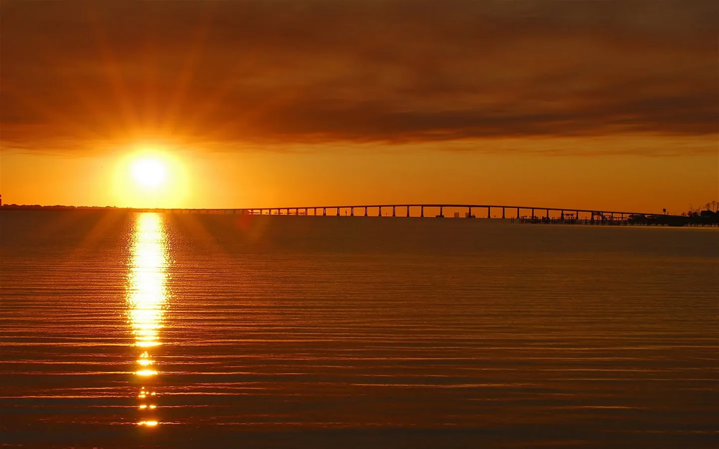 A bright sun sets over a calm ocean, casting a golden reflection on the water, with a long bridge in the distance.