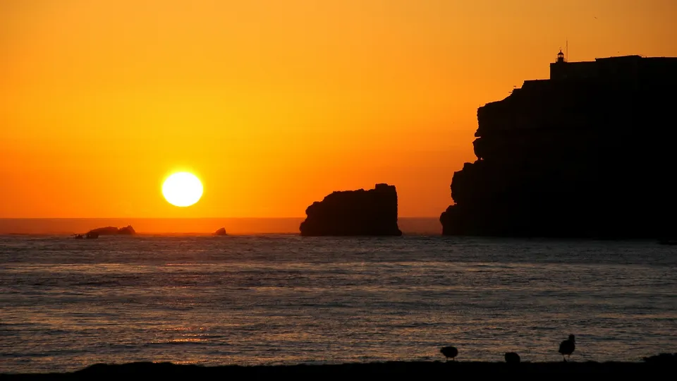 A bright sun sets over the ocean, casting a warm orange glow on the sky and water. Silhouetted cliffs and rocks are visible.