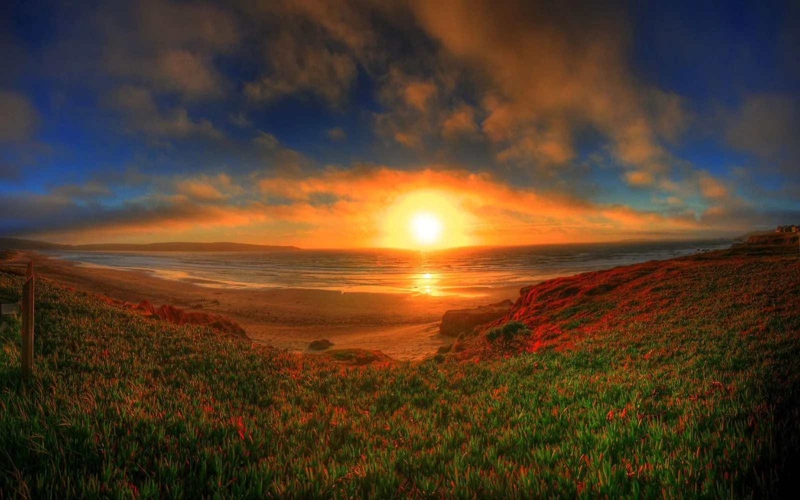 A bright sun sets over the ocean, casting golden light on a beach and grassy cliffs.