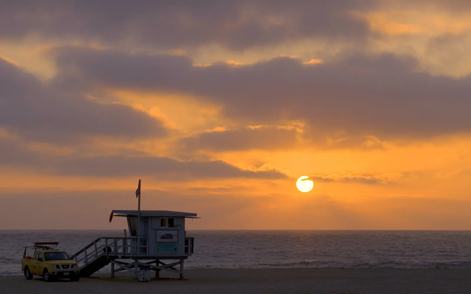 A lifeguard tower and truck on a beach at sunset with the sun setting over the ocean.