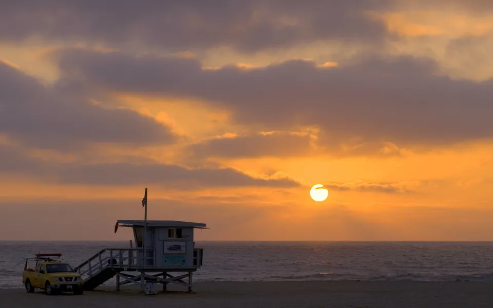 A lifeguard tower and truck on a beach at sunset with the sun setting over the ocean.
