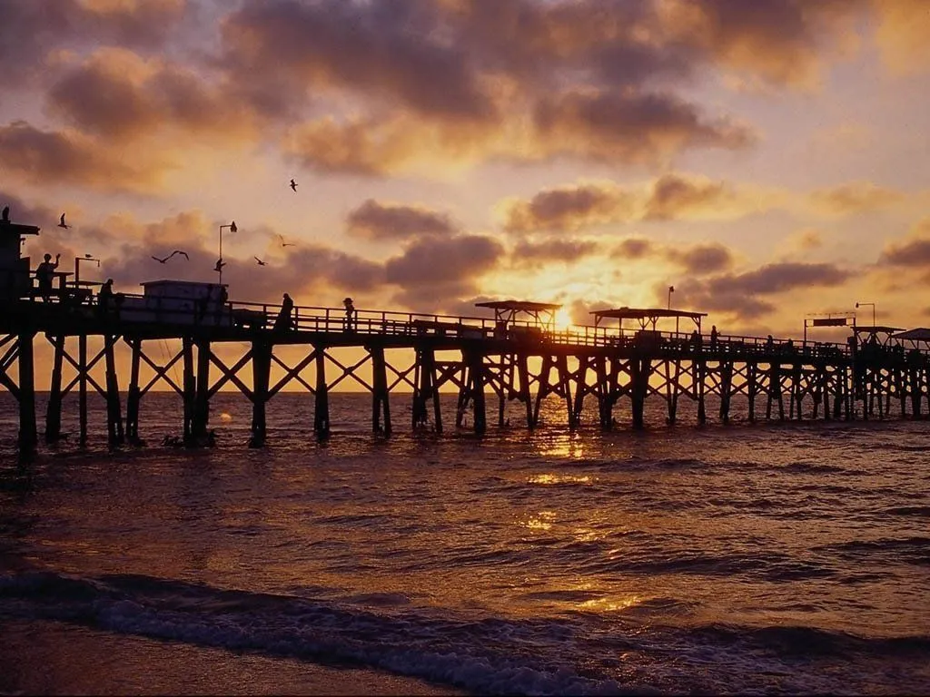 A silhouetted wooden pier stretches across the ocean during a vibrant sunset with clouds.