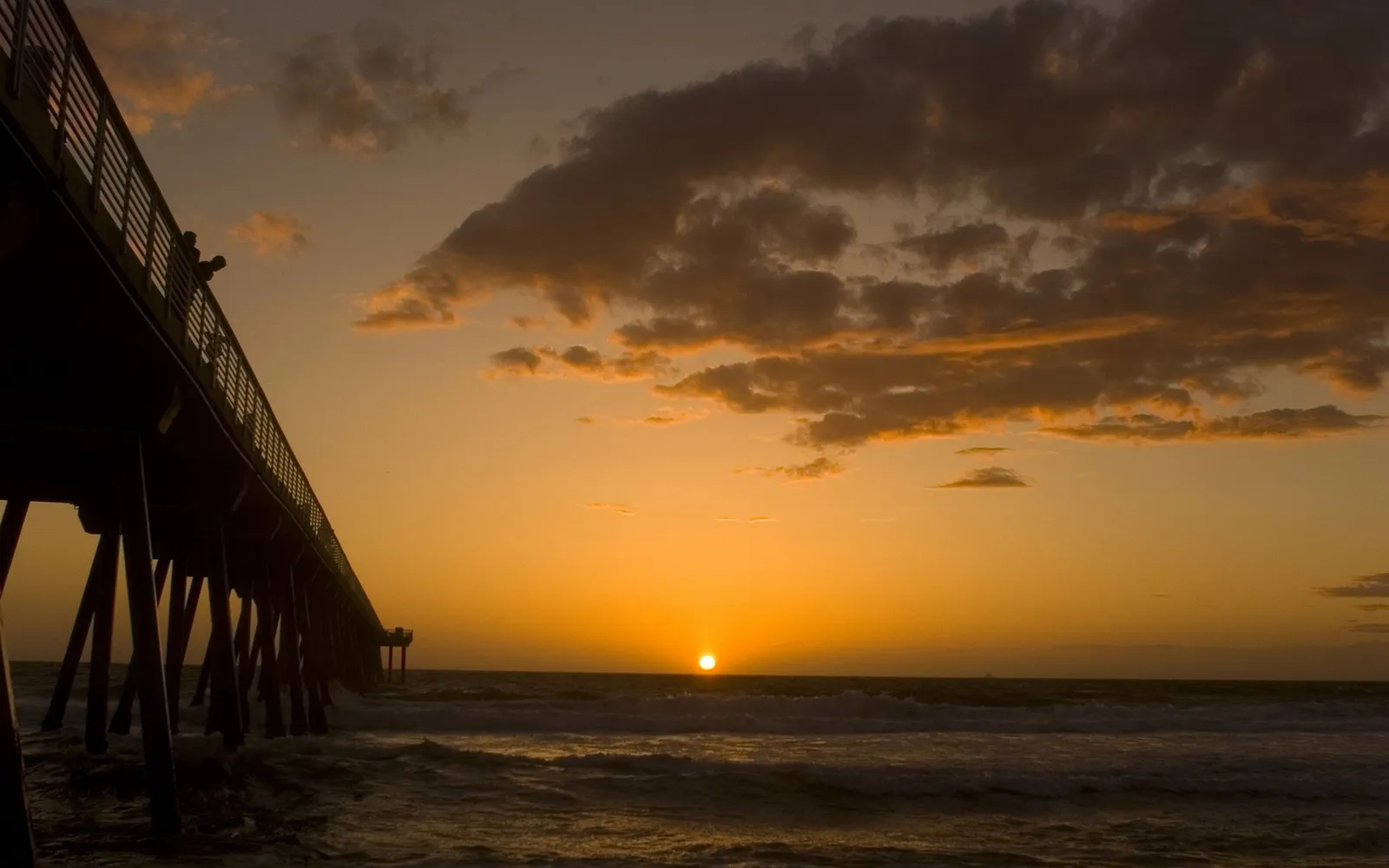 A pier extends over the ocean as the sun sets, casting a warm glow on the clouds and water.