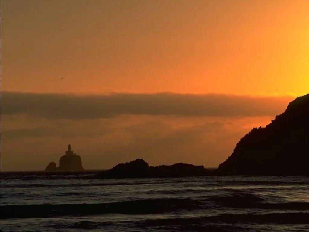 A silhouetted lighthouse sits on a rocky island in the ocean during a vibrant orange sunset.