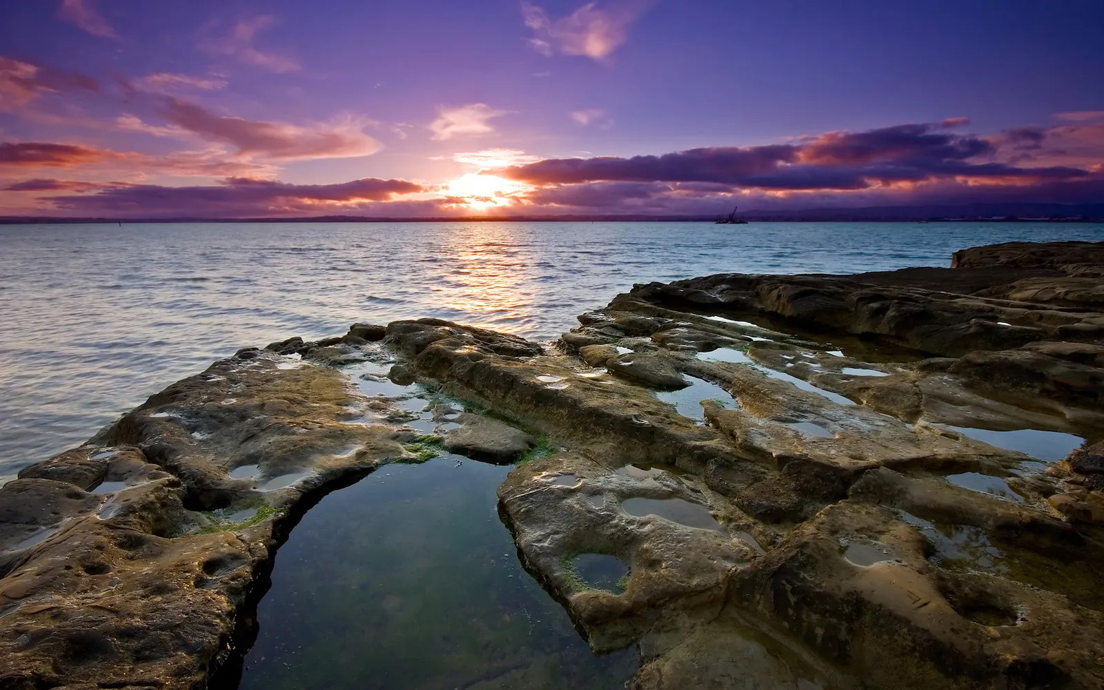 A dramatic sunset over the ocean with purple and orange clouds, reflecting on the water and wet rocks.