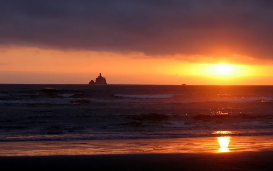 A dramatic sunset over the ocean with waves crashing on the shore and a sea stack in the distance.