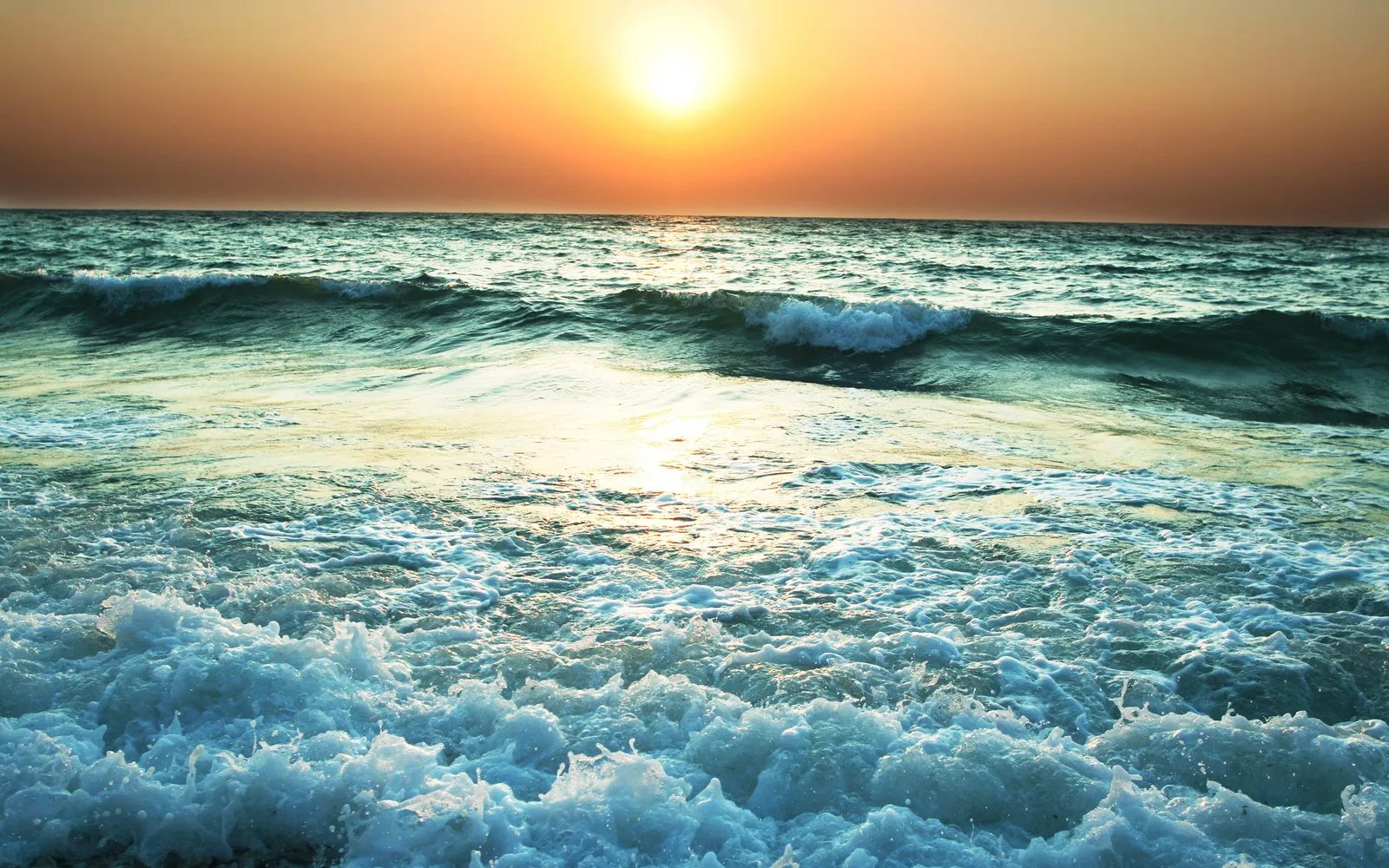 The sun sets over the ocean, casting a golden glow on the water as waves crash on the shore.
