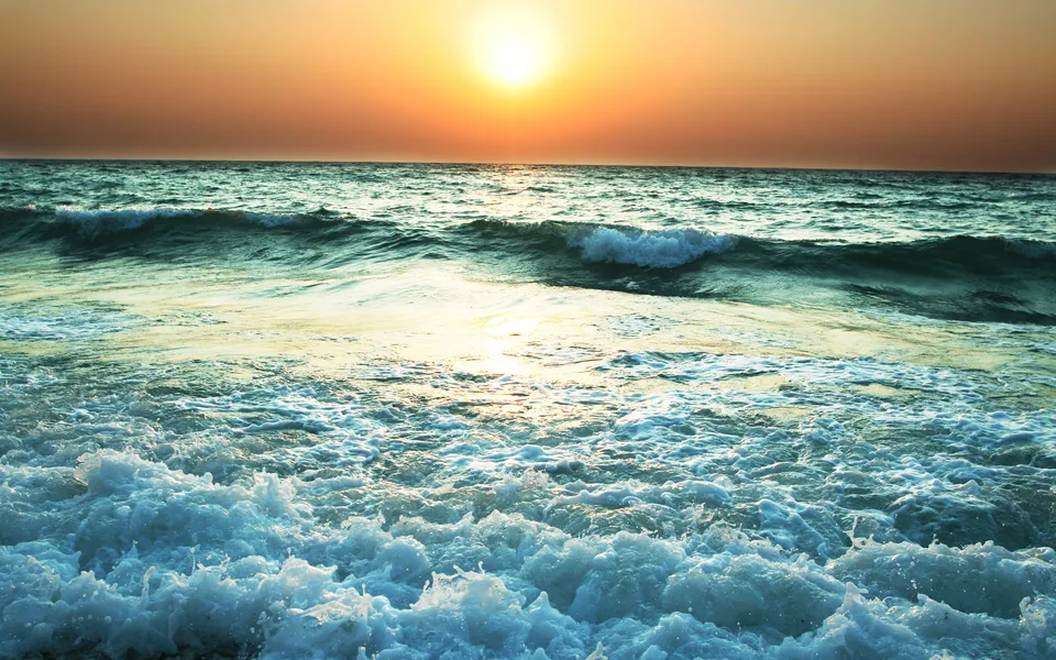 The sun sets over the ocean, casting a golden glow on the water as waves crash on the shore.