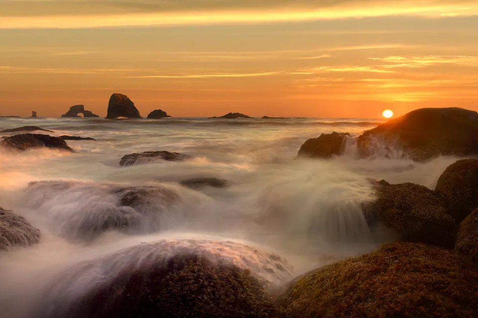 A serene sunset over a rocky coastline with misty waves crashing against the shore.