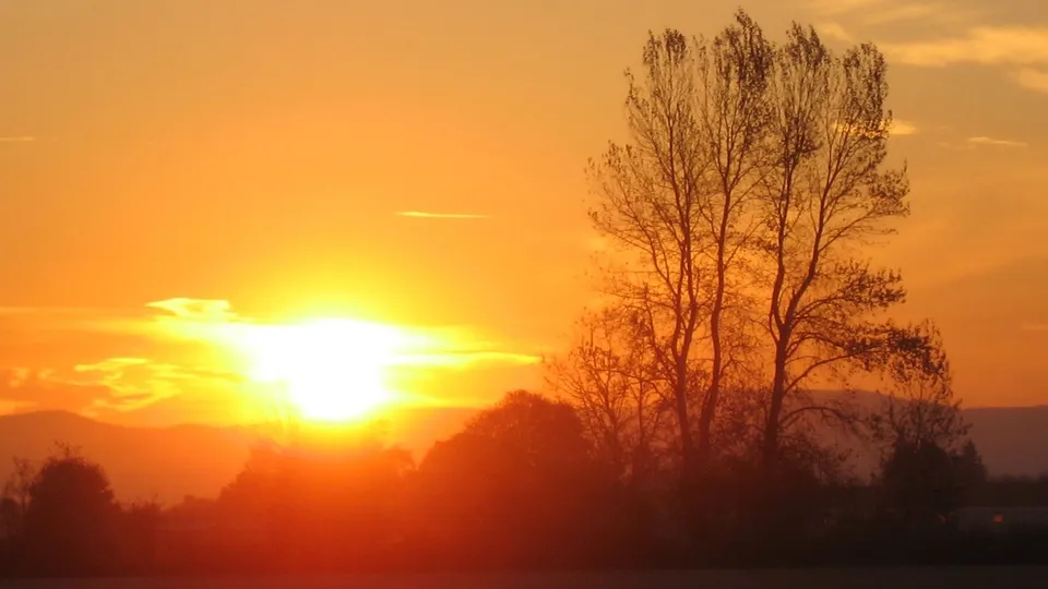 A bright sun sets behind distant hills, casting a warm golden glow over silhouetted trees and clouds.
