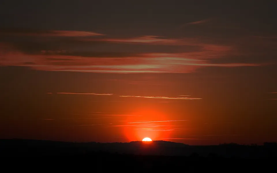The sun is setting behind a silhouetted hill, casting a vibrant orange glow across the sky with wispy clouds.