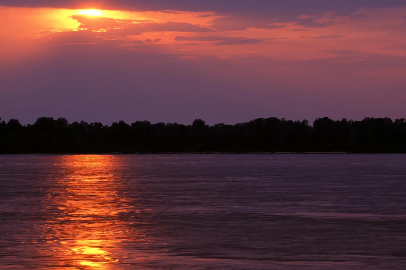 A vibrant sunset casts an orange glow on the Mississippi River, with silhouetted trees on the far bank.