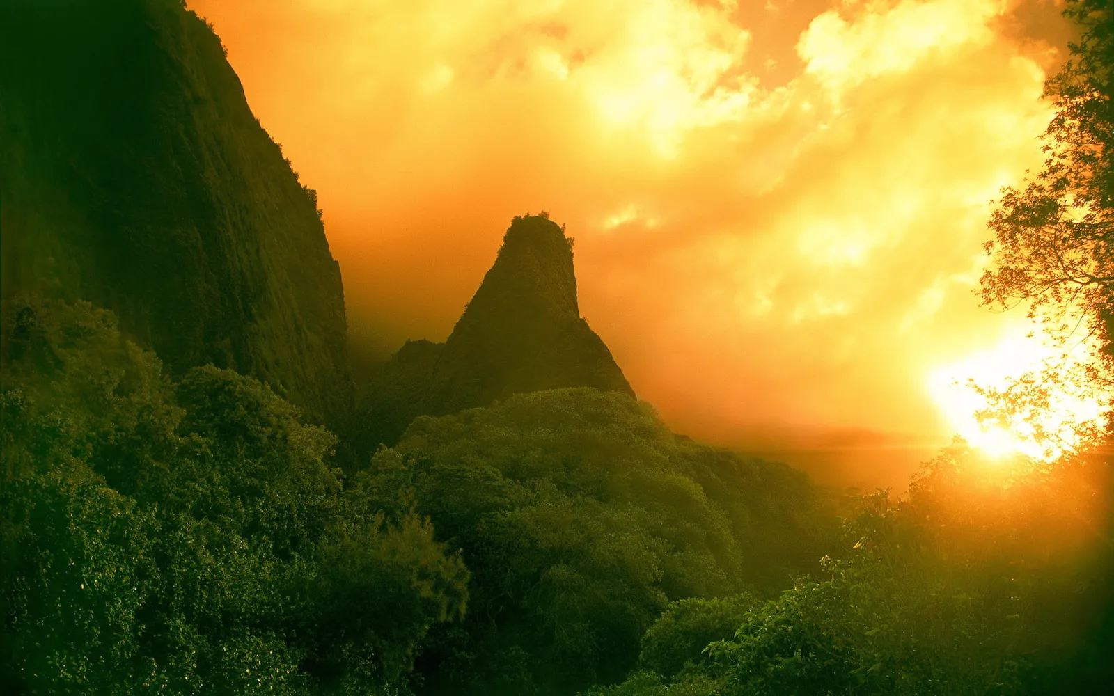 A jagged mountain peak is silhouetted against a vibrant orange sky with dramatic clouds during sunset.