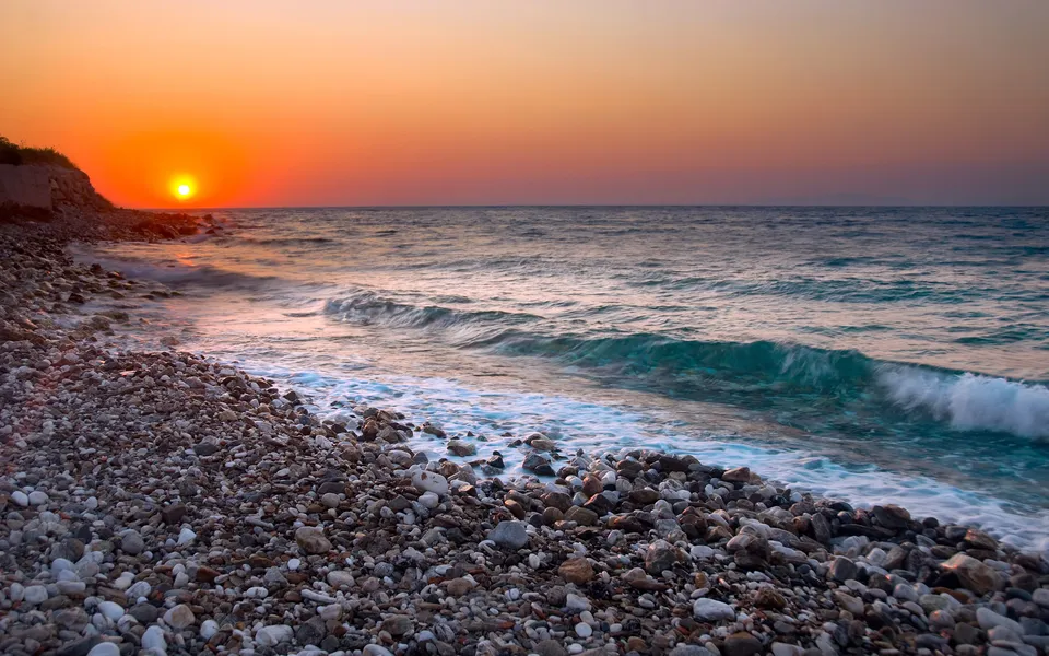 A vibrant sunset casts golden light over a pebble beach with gentle waves lapping the shore.