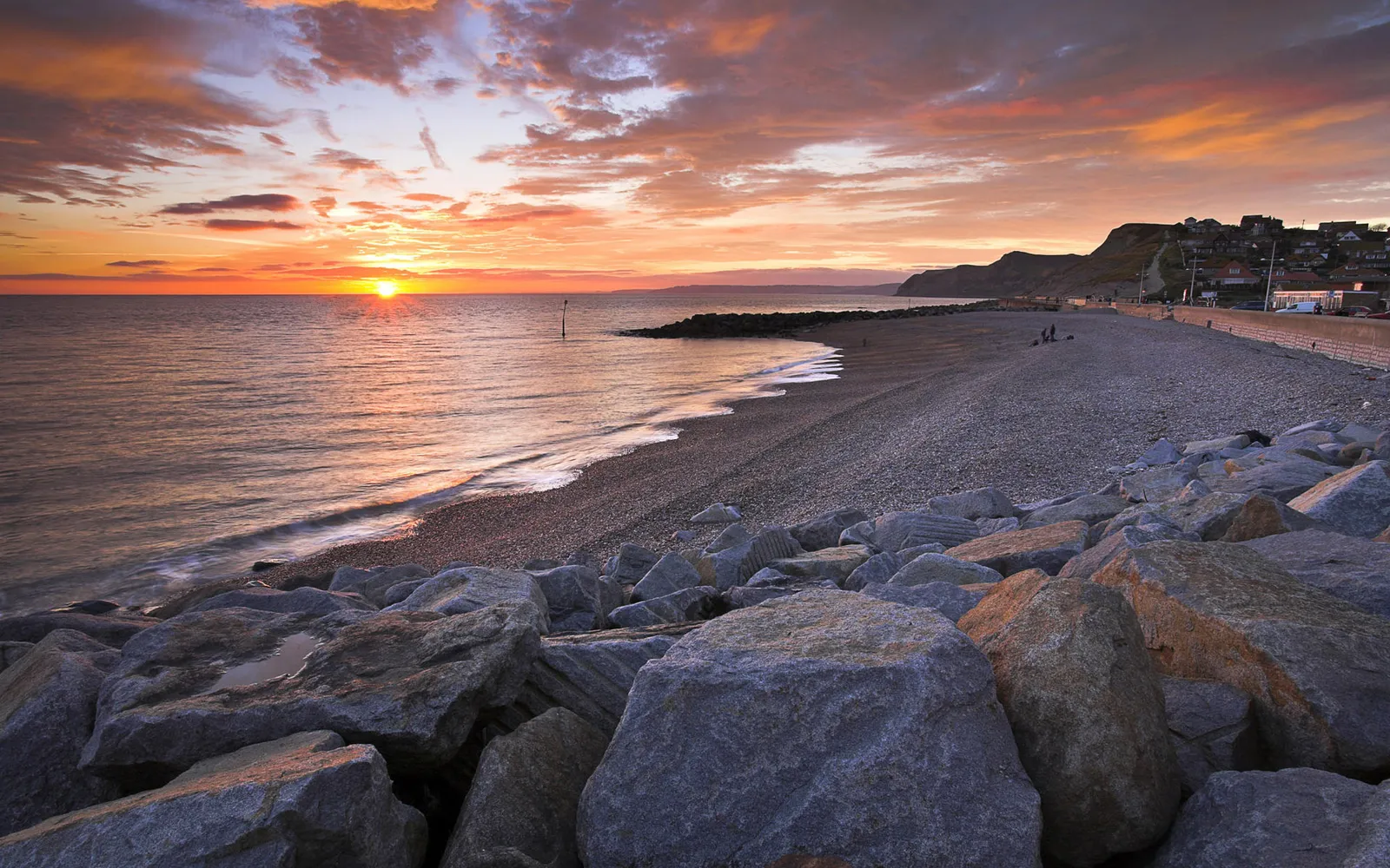 A vibrant sunset over a pebble beach with large rocks in the foreground and a town on the hillside.