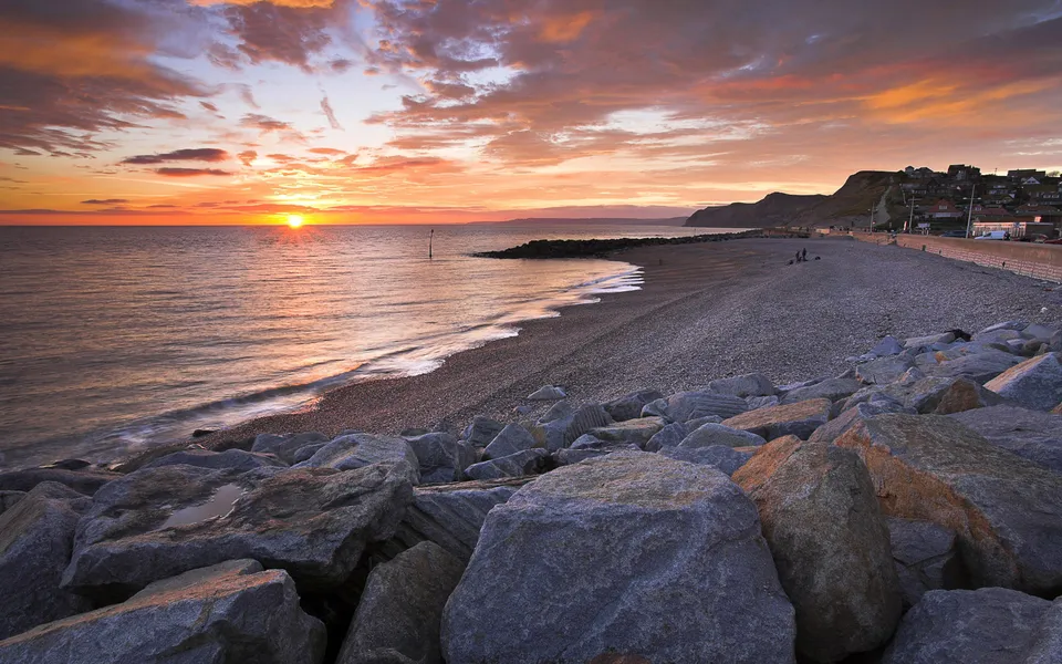 A vibrant sunset over a pebble beach with large rocks in the foreground and a town on the hillside.