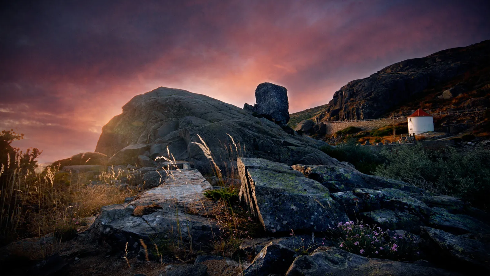 A rugged mountain landscape at sunset, with a small white building visible on the hillside.