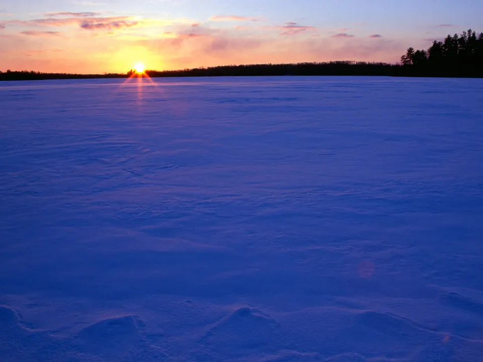 The sun sets over a vast, snow-covered field with a dark treeline in the distance.