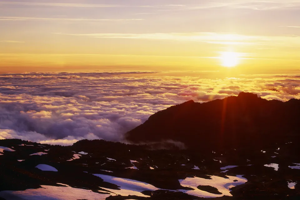 A golden sunset illuminates a vast sea of clouds below silhouetted mountain peaks and snow-dusted terrain.