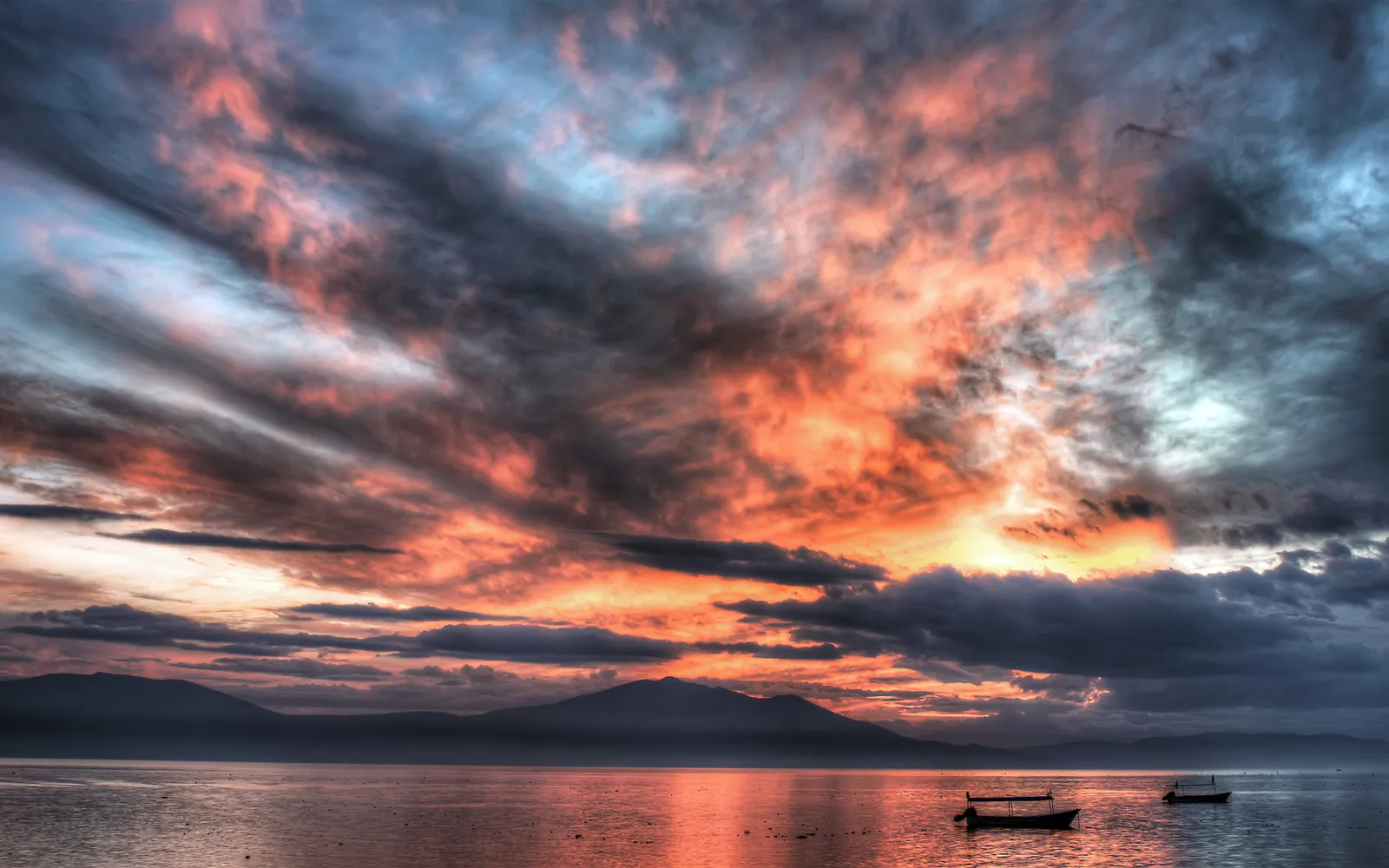 A dramatic sunset with fiery orange and pink clouds over a calm lake with distant mountains and two small boats.