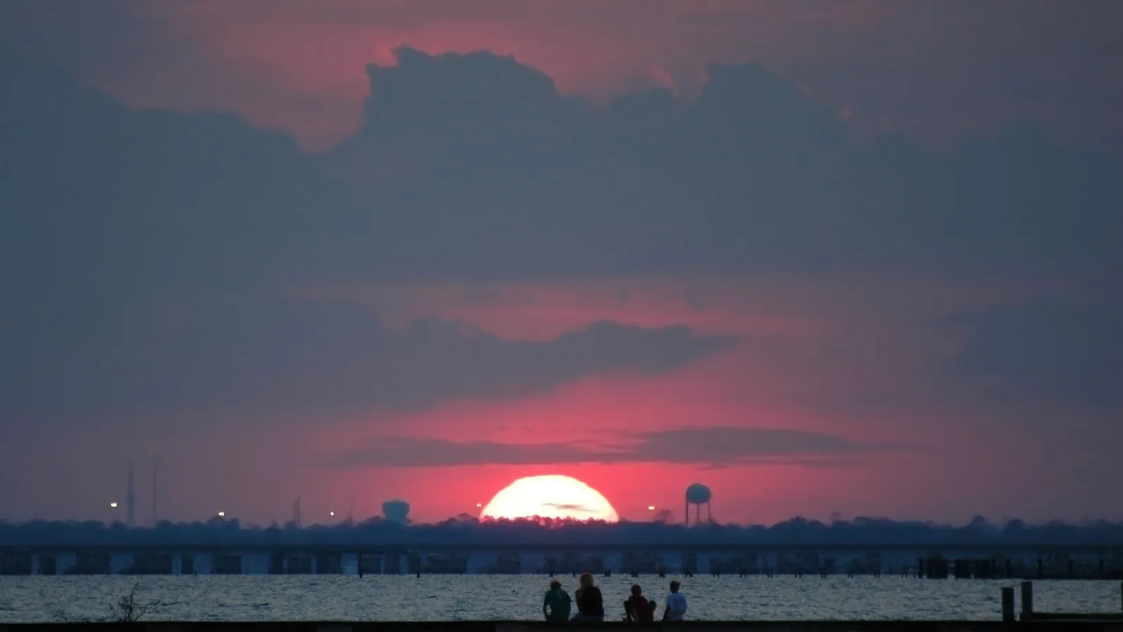 A large, bright sun sets over a body of water, casting a pink and orange glow on the clouds. Silhouetted figures sit on a pier in the foreground.