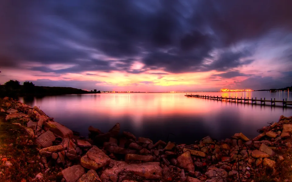 A dramatic sunset with colorful clouds over a calm body of water, featuring a long pier and rocky foreground.
