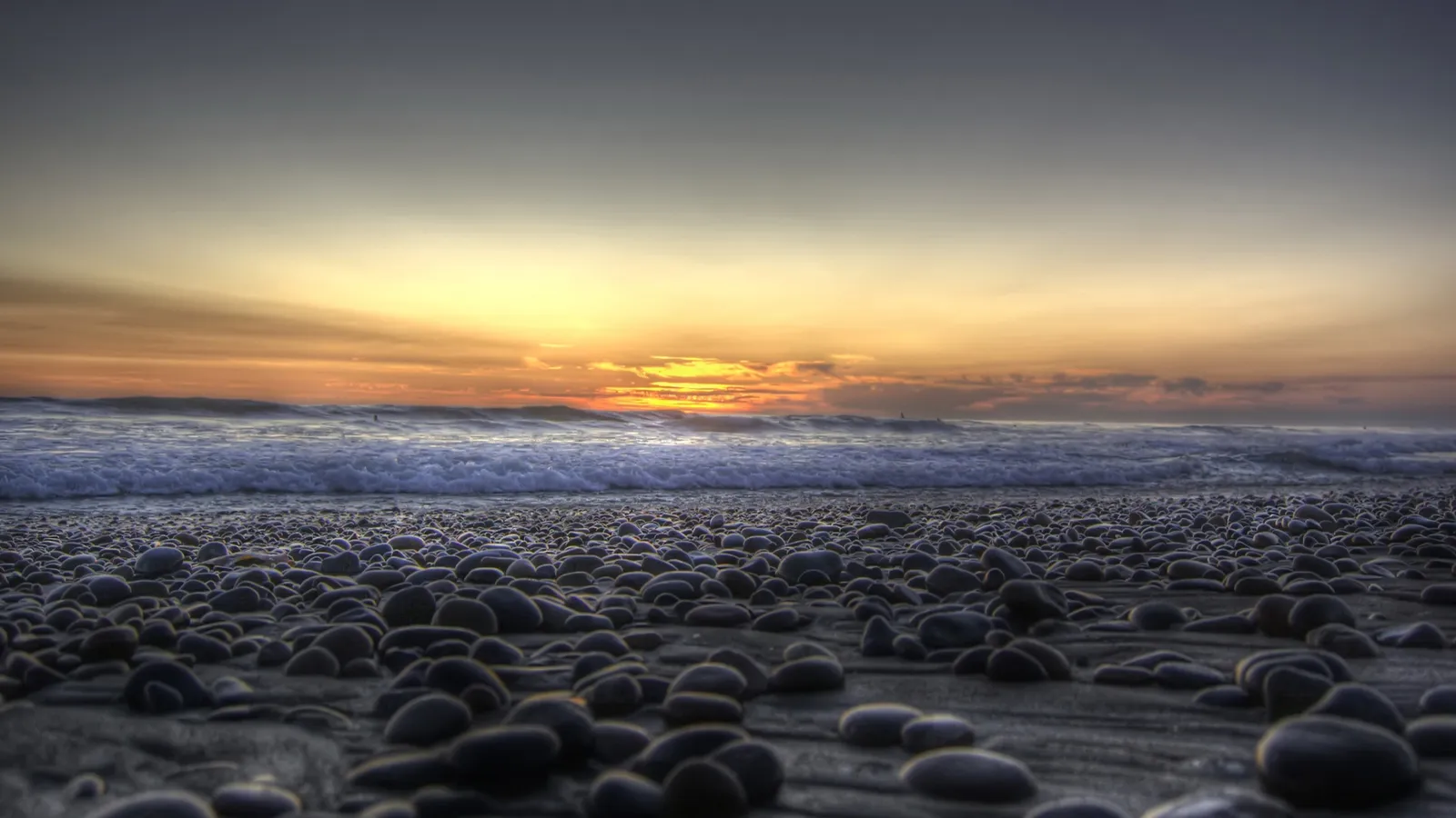 A pebble beach with waves rolling in during a colorful sunset.