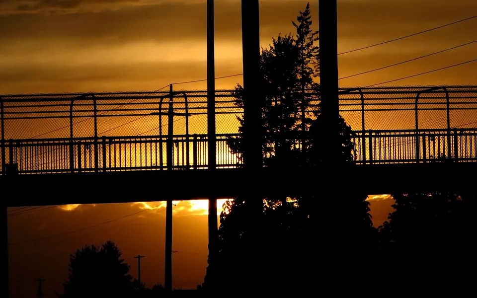 A pedestrian bridge is silhouetted against a vibrant orange and yellow sunset sky.
