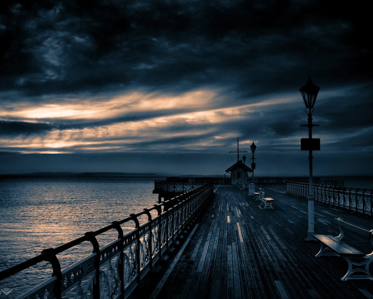 A wooden pier stretches out over the ocean under a dramatic, cloudy sky at sunset.
