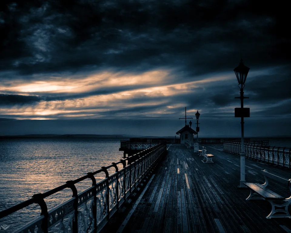 A wooden pier stretches out over the ocean under a dramatic, cloudy sky at sunset.