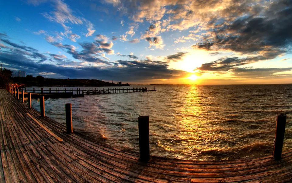 A wooden pier extends into a body of water as the sun sets, casting a golden glow.