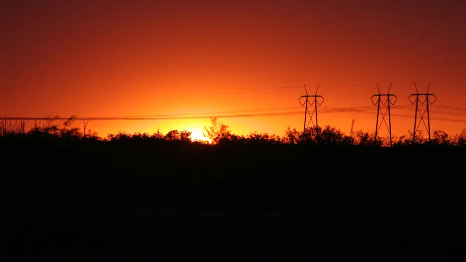 A vibrant orange and red sunset behind a silhouetted line of trees and power line towers.