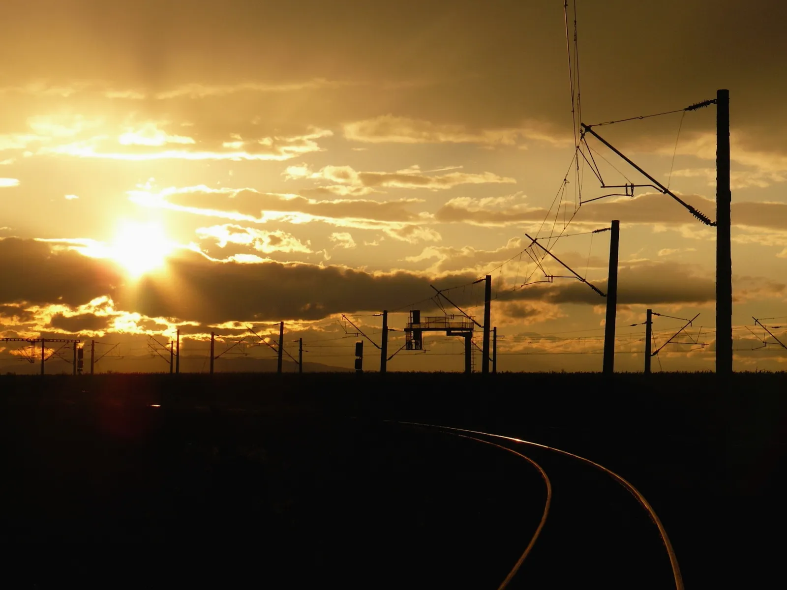 A dramatic sunset with golden sunbeams shining through clouds over silhouetted railway tracks and power line poles.