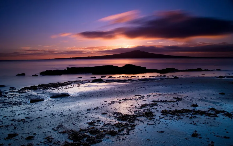A dramatic sunset over Rangitoto Island, viewed from a rocky beach with calm water.
