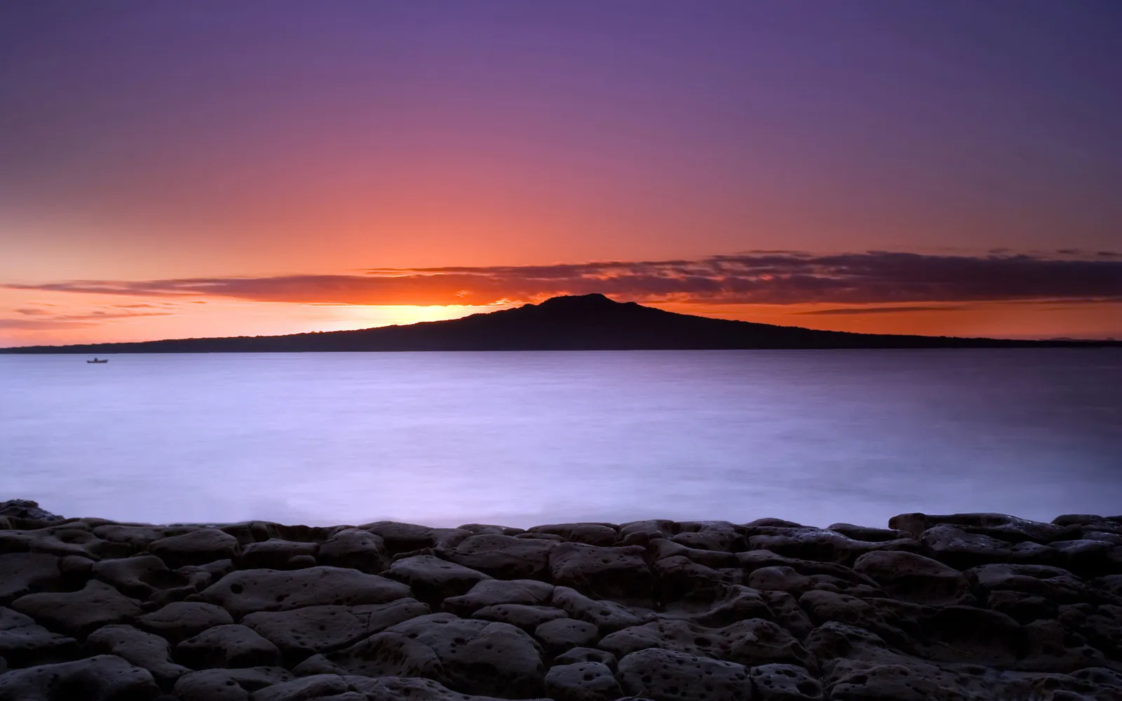 A silhouette of Rangitoto Island is visible across a calm sea during a vibrant sunset.