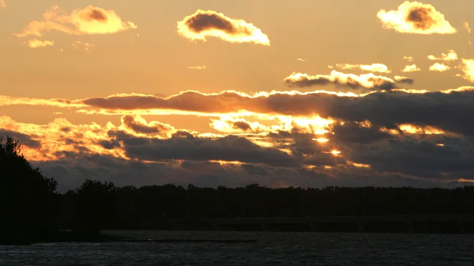 The sun sets behind dark clouds, casting golden rays over a silhouetted treeline and water.