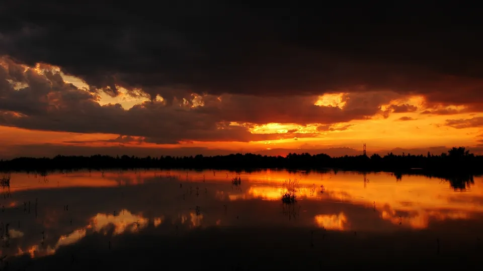 A dramatic sunset with fiery orange clouds reflected in a still body of water, silhouetting a treeline.