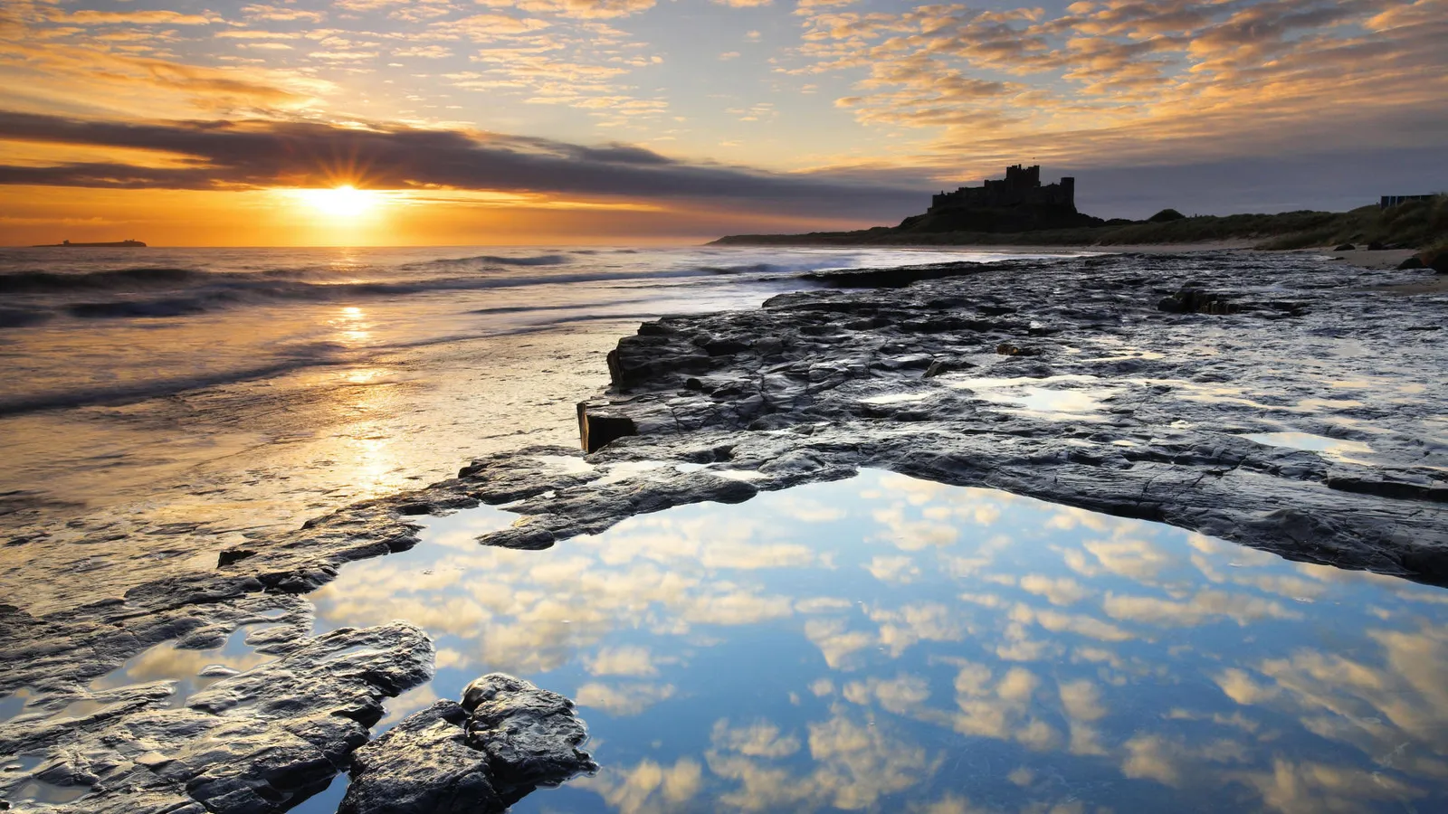 A dramatic sunset over a rocky coastline with a castle in the distance and a reflection in a tidal pool.